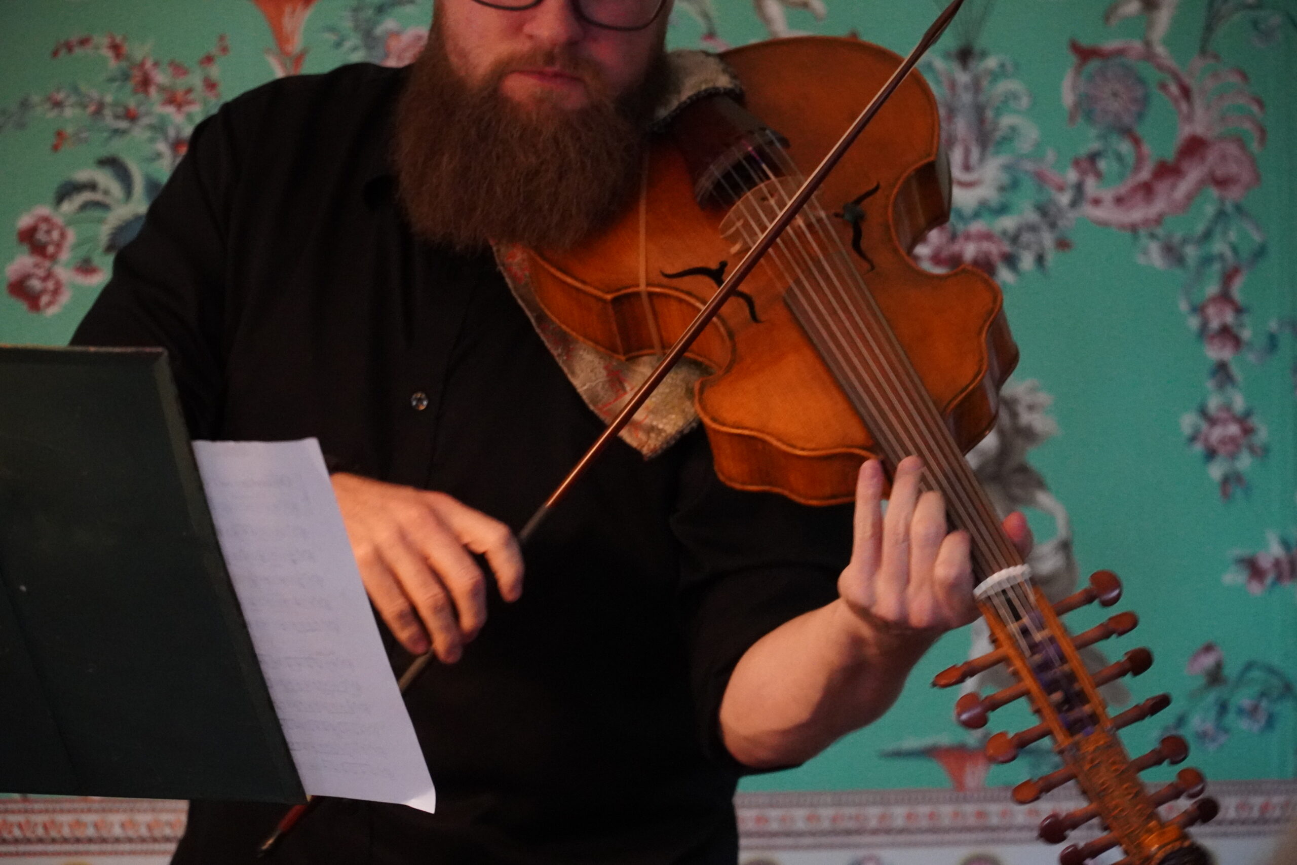 Man in black clothing stands in front of seafoam wallpaper holding a violin in front of a music stand.