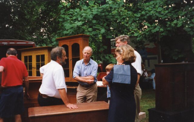 three men, a woman, and child talk with wooden furniture around them.