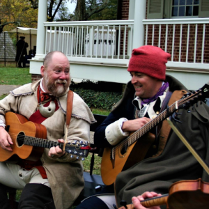 Two musicians in 18th century clothing play guitars in front of a brick historic house